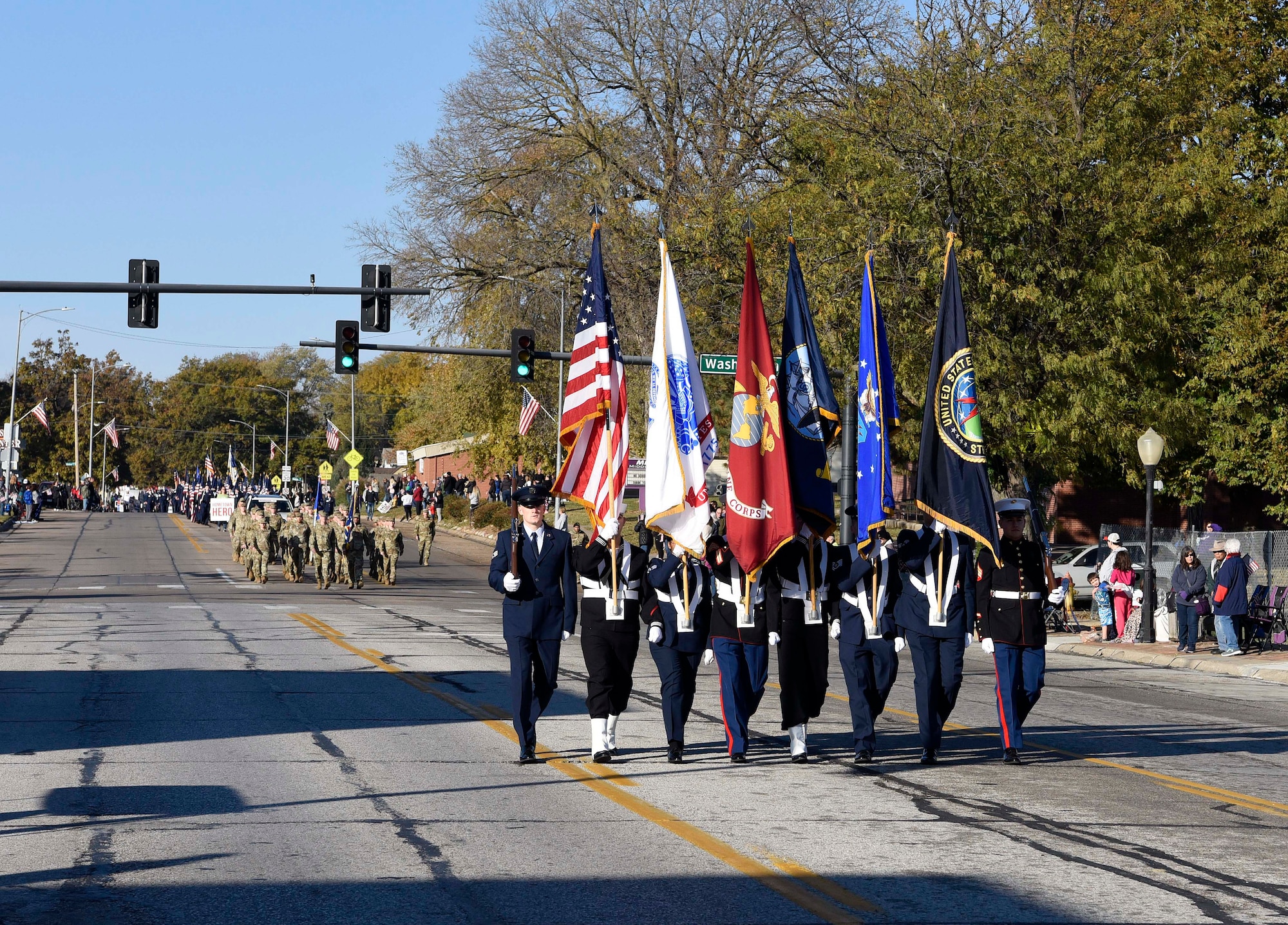 A color guard with 8 service members in dress uniforms from different military branches marches down the middle of a street.