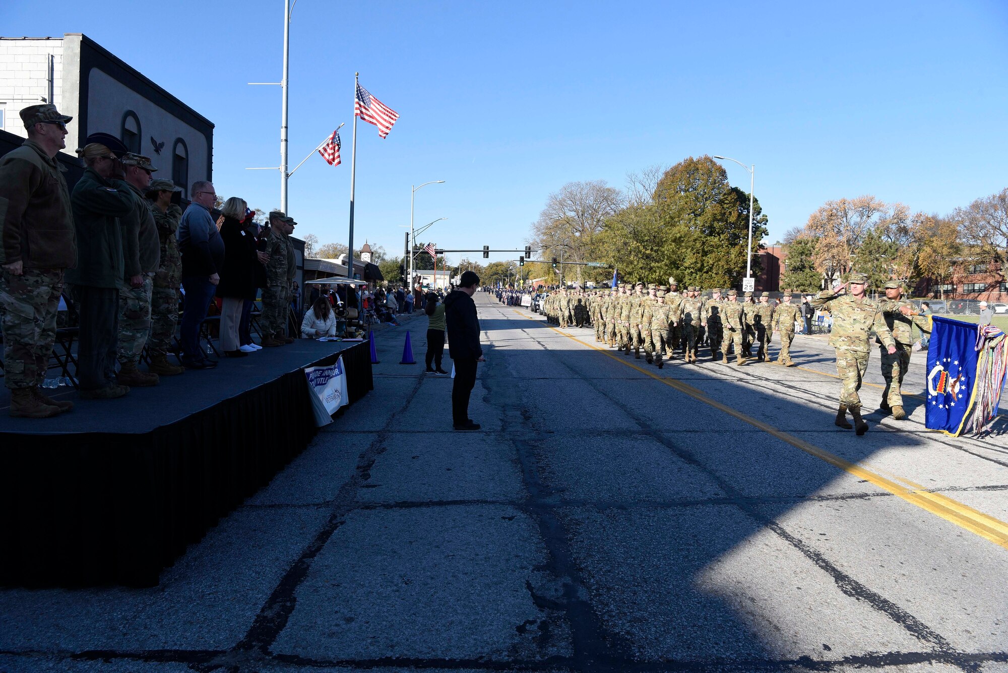 Military members march in ranks and salute in front of military and civilian officials on a raised stage.