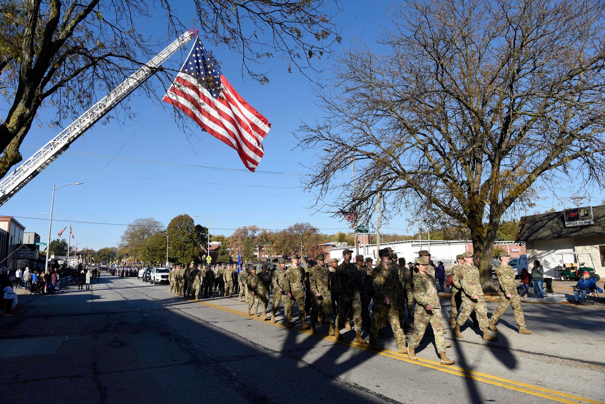 Military members in camouflage uniforms march in ranks down the middle of the street under a giant U.S. flag hung at the top of a fire truck ladder.