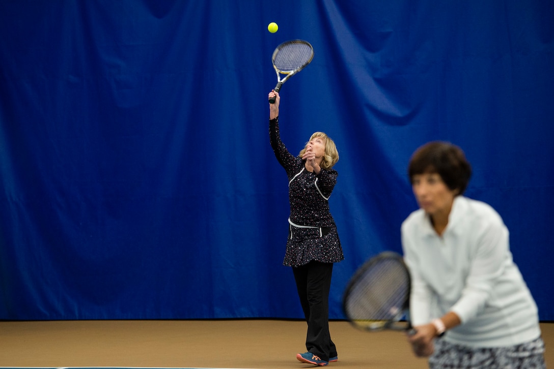 Kathi Malina, serves up a tennis ball while her partner Annette Oakes waits for the ball to be returned while playing tennis inside the base Tennis Club, Nov 1, 2021, at Wright-Patterson Air Force Base, Ohio. The club offers 4 LED lit courts to play on and several other amenities for those who need to practice. (U.S. Air Force photo by Wesley Farnsworth)