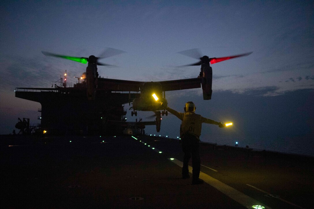 A sailor signals to an aircraft aboard a ship at sea in the dark illuminated by yellow, green and red lights.