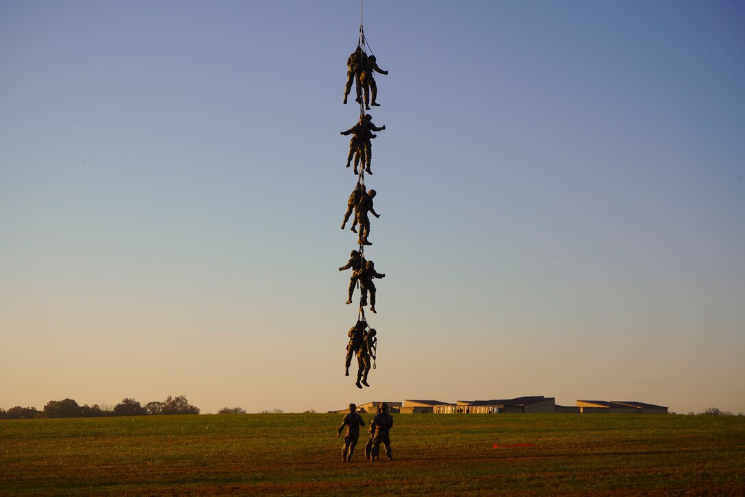 Ten soldiers lift off the ground on a rope attached to helicopter as fellow soldiers watch from below.