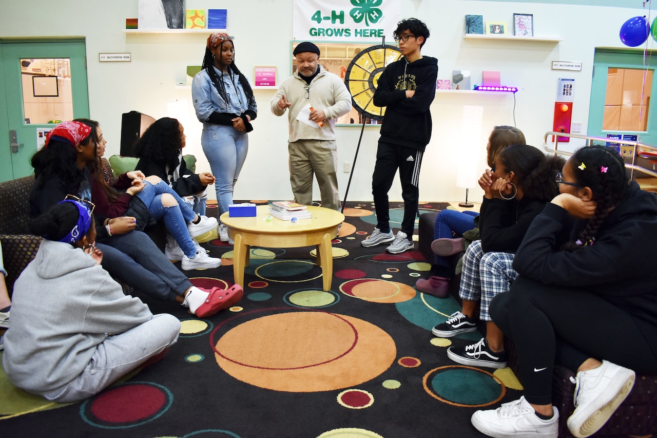 Several seated teens look at a counselor and two other teens who are standing at the front of a room.