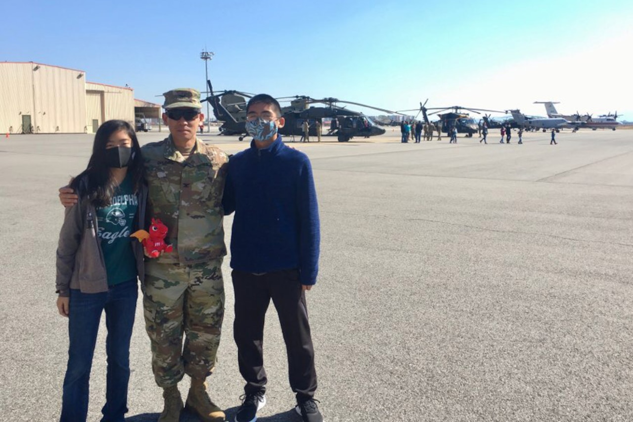A soldier puts his arms around a teenage girl and boy for a photo on tarmac. Helicopters can be seen behind them.