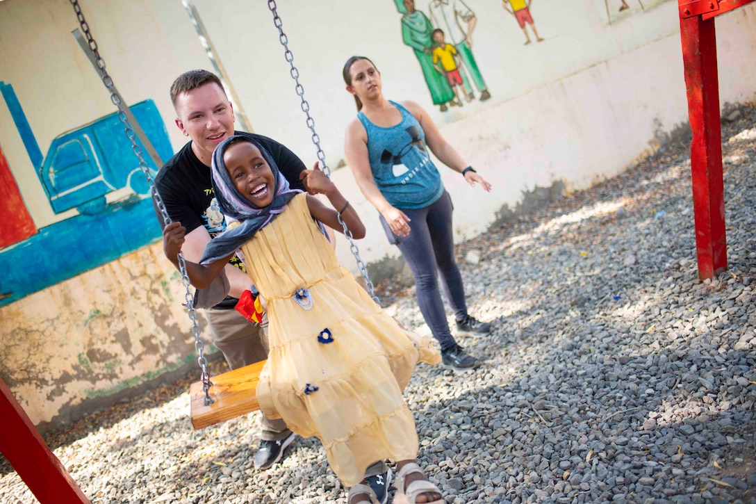 A sailor pushes a child on a swing as a woman stands behind.