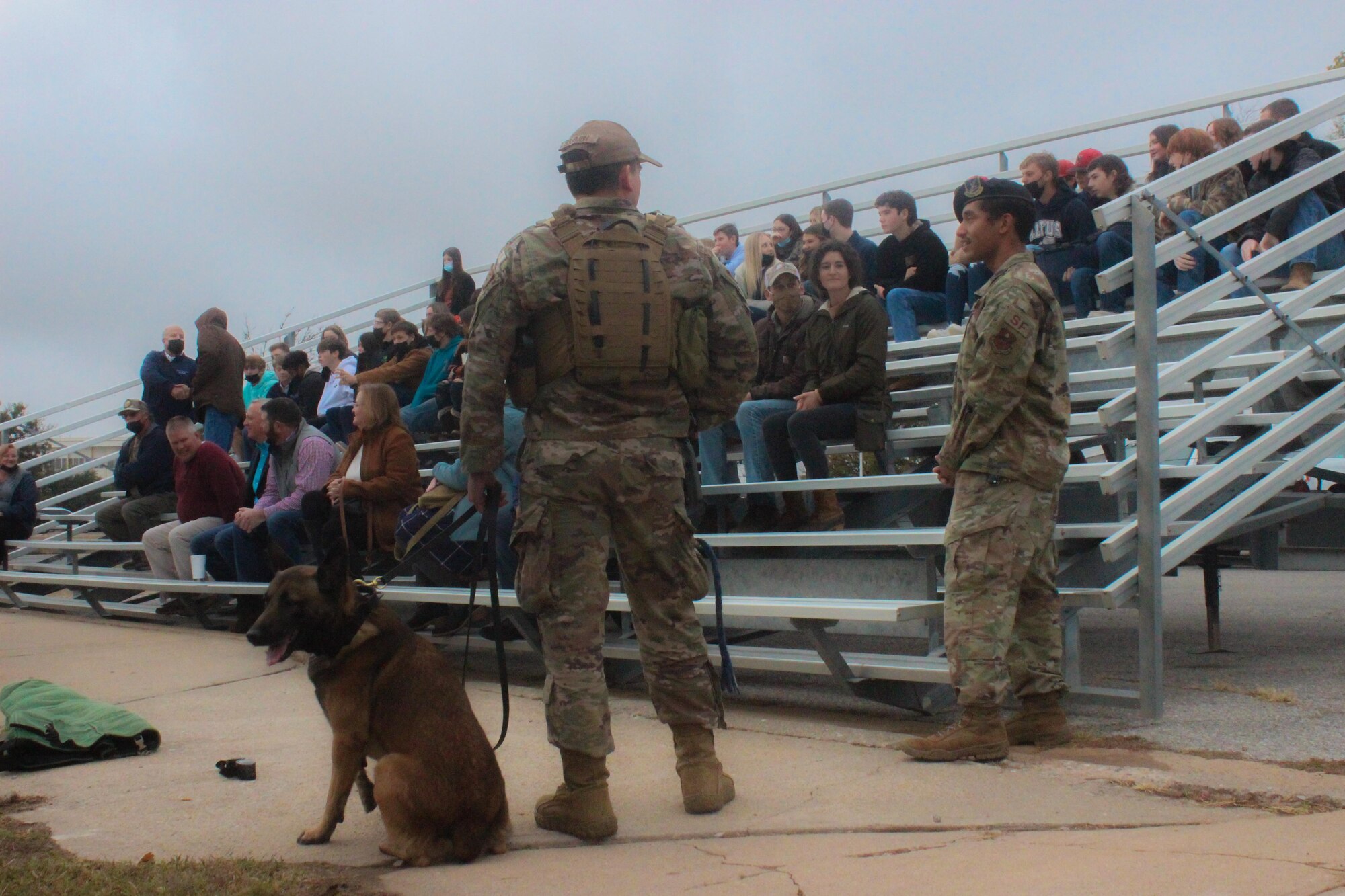 Two men and a dog stand near bleachers full of students