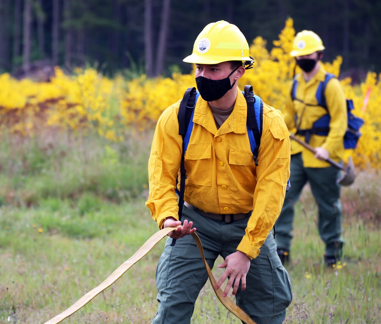 Soldier wearing yellow fire resistant uniform plays out a hose. Behind him, another firefighter stands ready with a shovel.