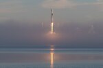 A rocket takes off, its contrail reflected in a nearby body of water.