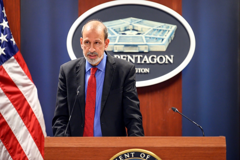 A man stands behind a lectern. Behind him is a wall-hanging with a picture of the Pentagon.