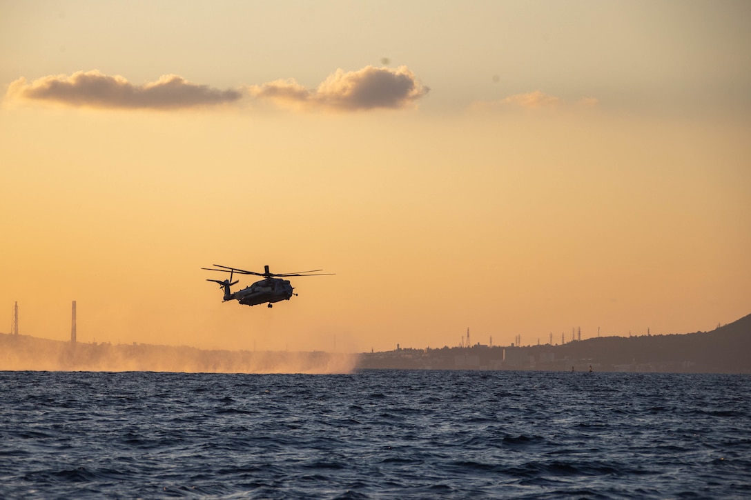 A helicopter flies just above the water at twilight.