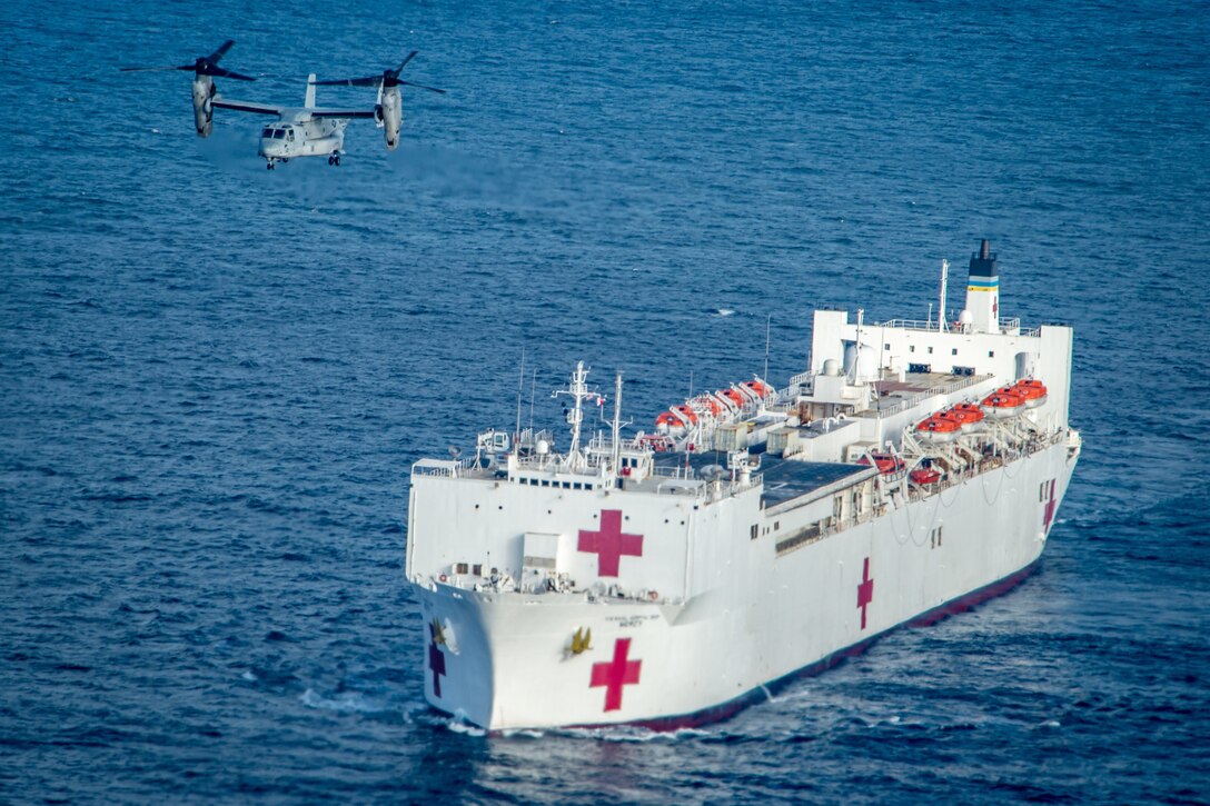 An Osprey aircraft flies above a military hospital ship in the ocean.