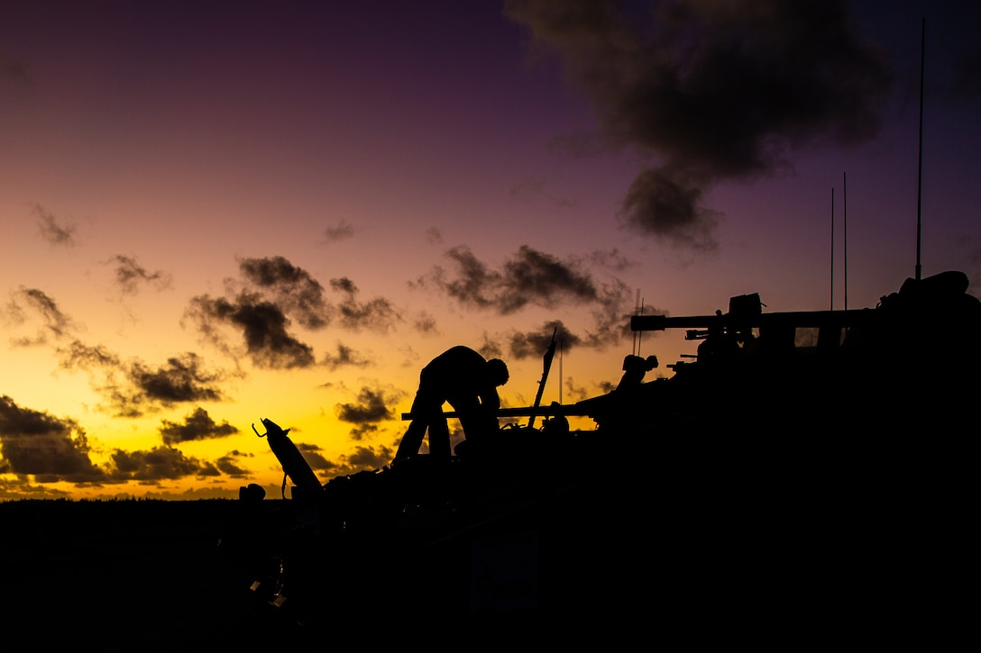 A Marine stands on a tank while parked next to another tank under a sunlit sky as seen in silhouette.