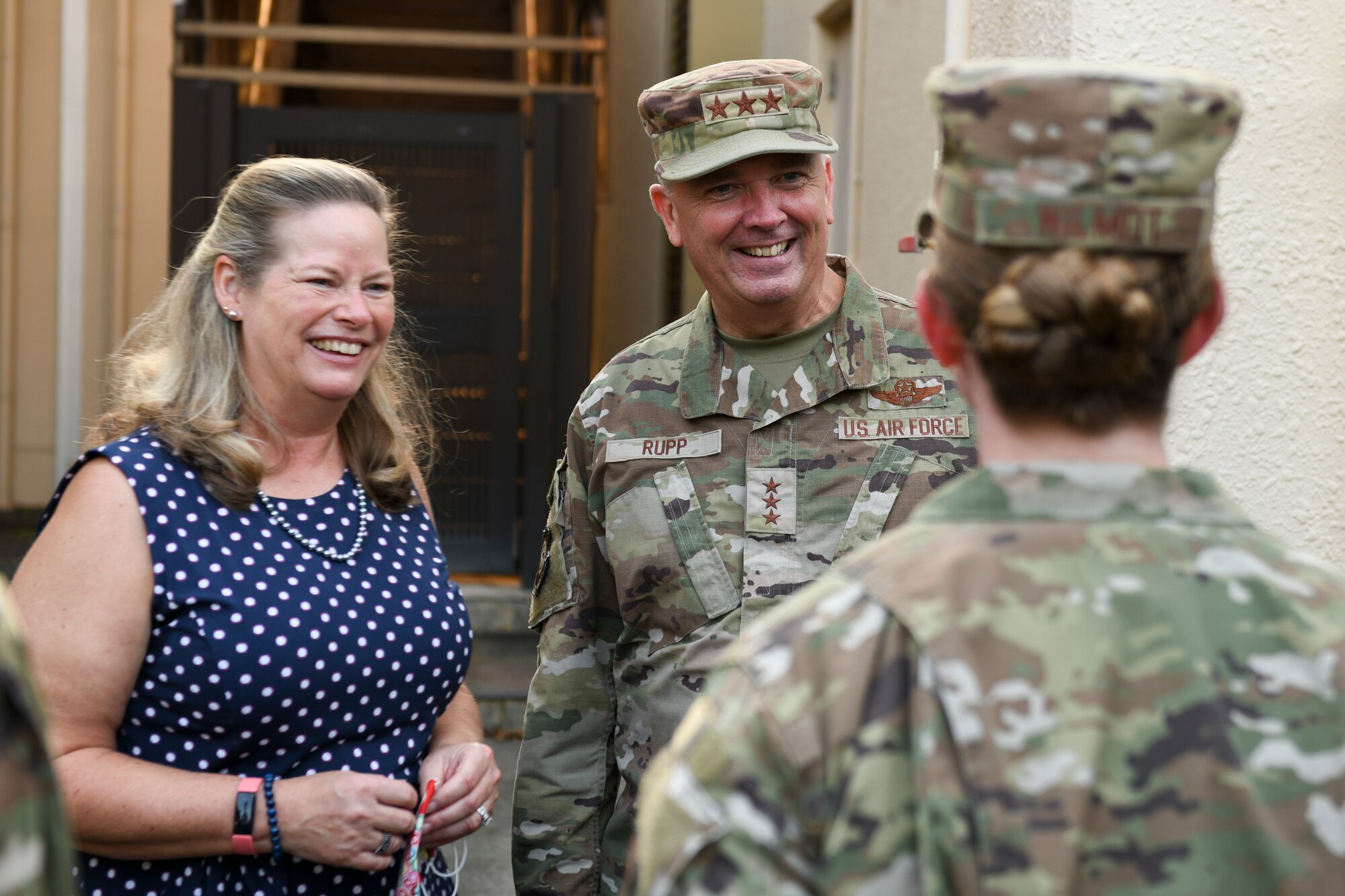 A 3-star general and his wife smile while talking to an Airman.