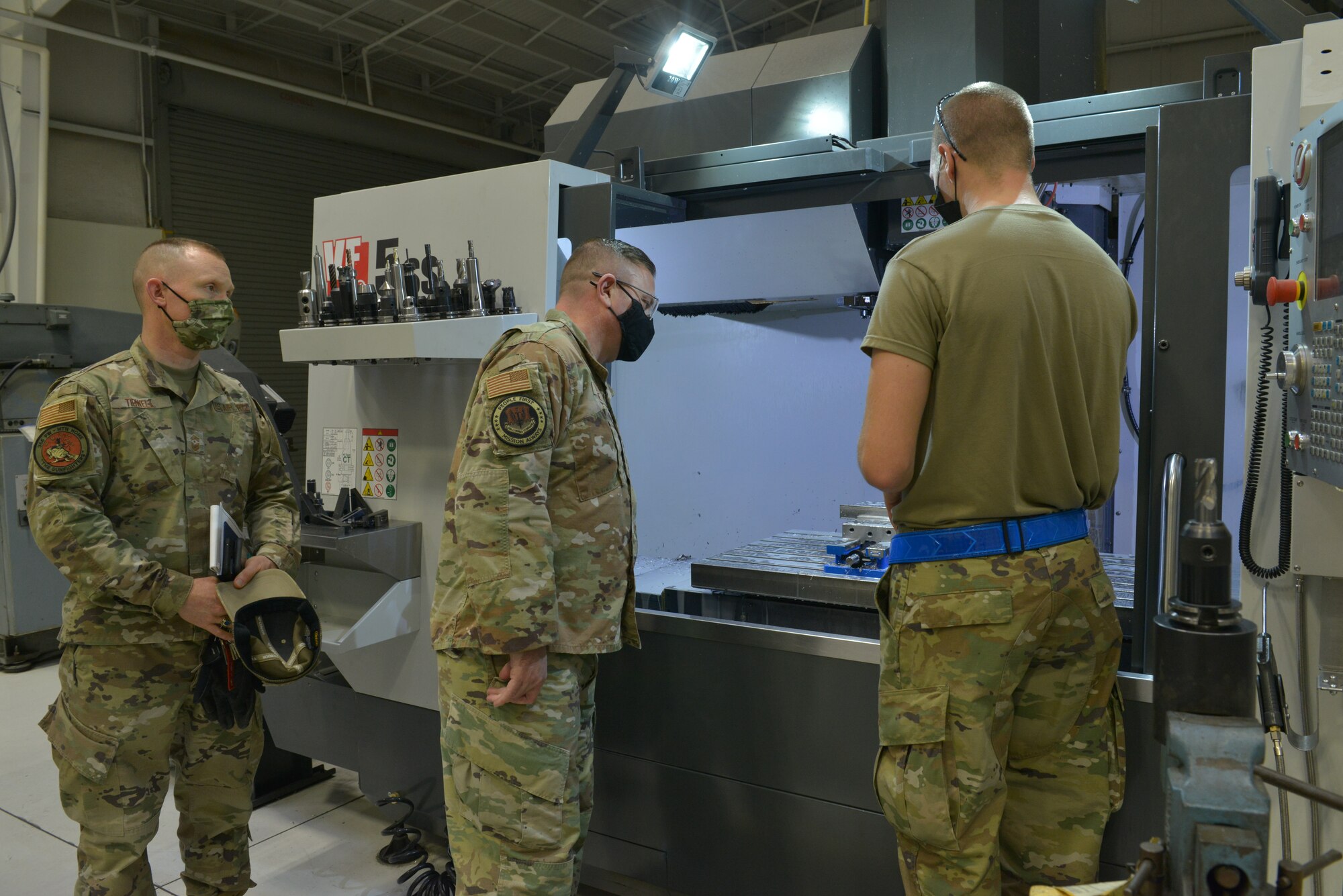U.S. Air Force Tech. Sgt. Issac McDonald, right,  366th Maintenance Squadron metals tech craftsman, briefs Command Chief Master Sgt. David Wade, Air Combat Command, and Chief Master Sgt. Joshua Tidwell, 366th Fighter Wing command chief, on the Computer Numeric Control (CNC) machine, on Mountain Home Air Force Base, Idaho, Nov. 9, 2021. This machine uses a cutter tool with careful calculations to coordinate data input into the machine, to make precision cuts to any block material that the Airmen place into the machine.