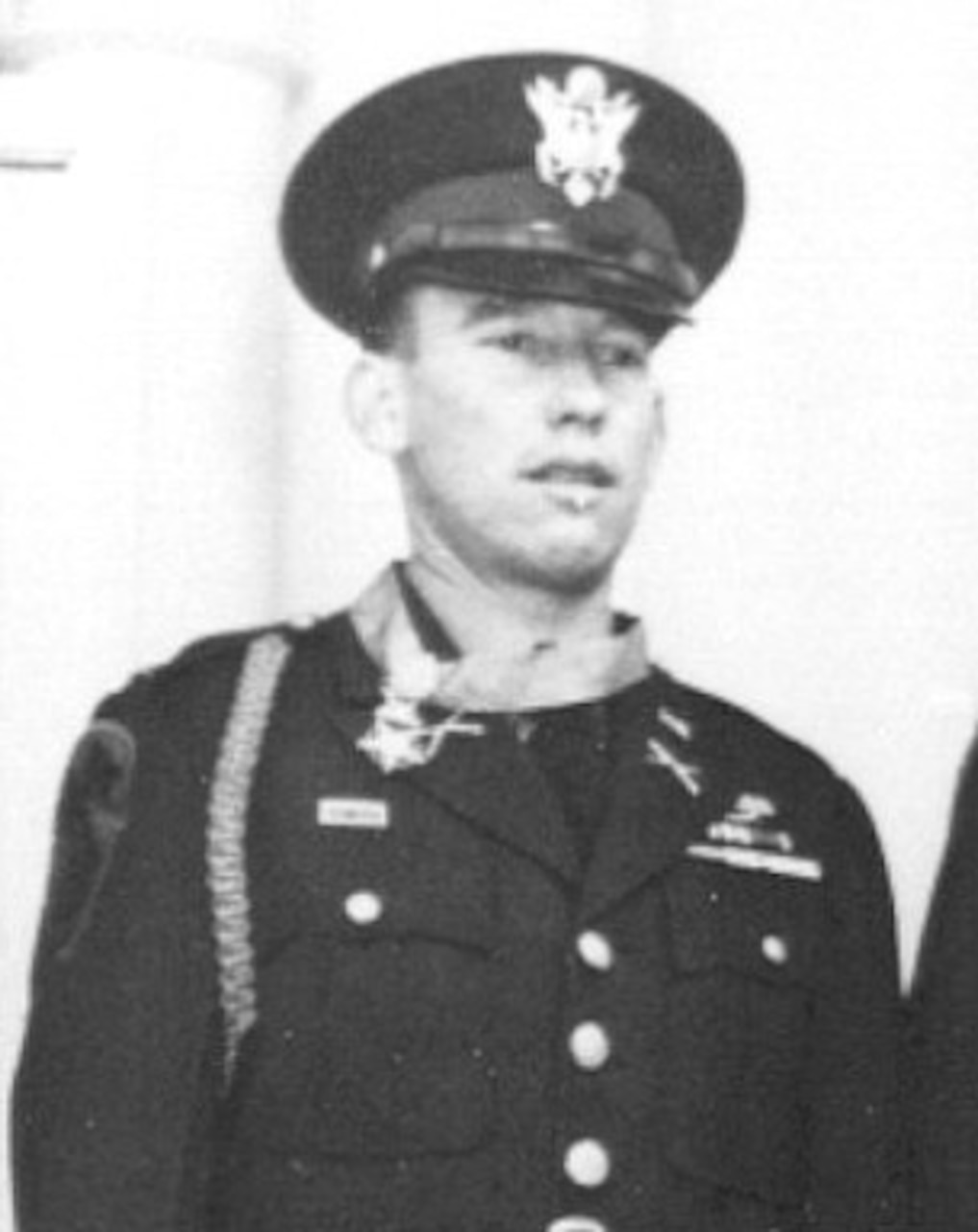 A man in dress uniform and cap wears a medal around his neck.
