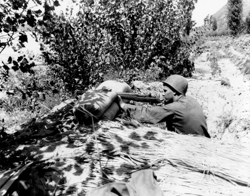 A soldier props a rifle on a sandbag near a hill and trees.