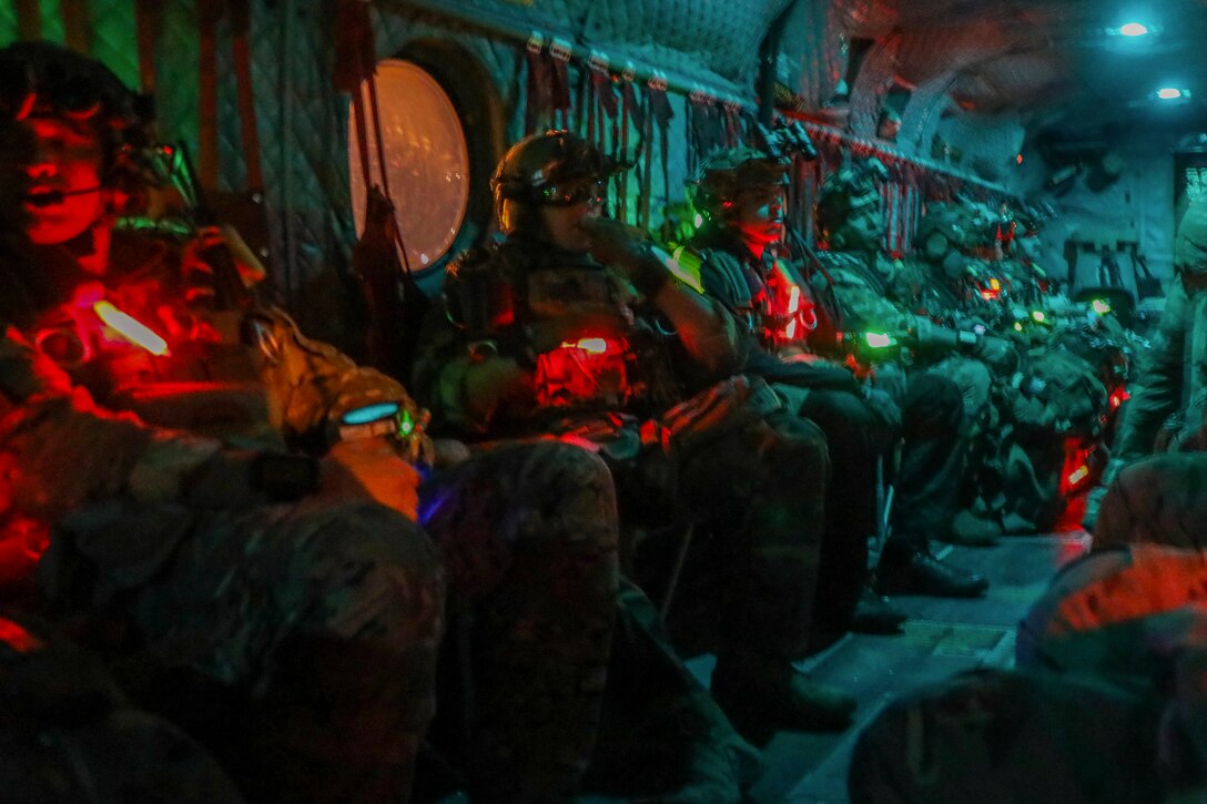 Soldiers sit next to one another on an aircraft in the dark illuminated by colorful lights.