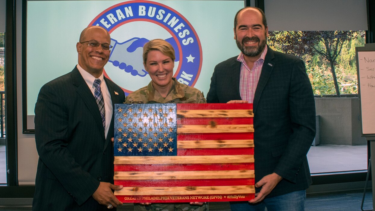 A woman in uniform poses between two men while holding a wooden American Flag.