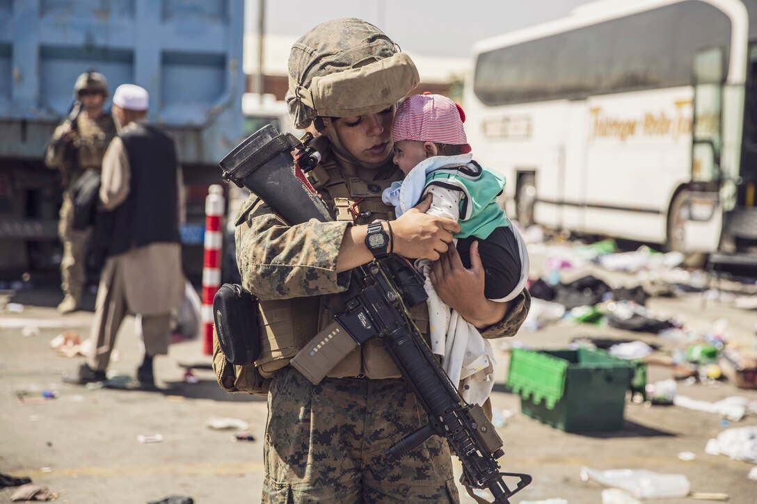 A Marine with a rifle strapped to her uniform holds an infant in a litter-strewn urban environment.
