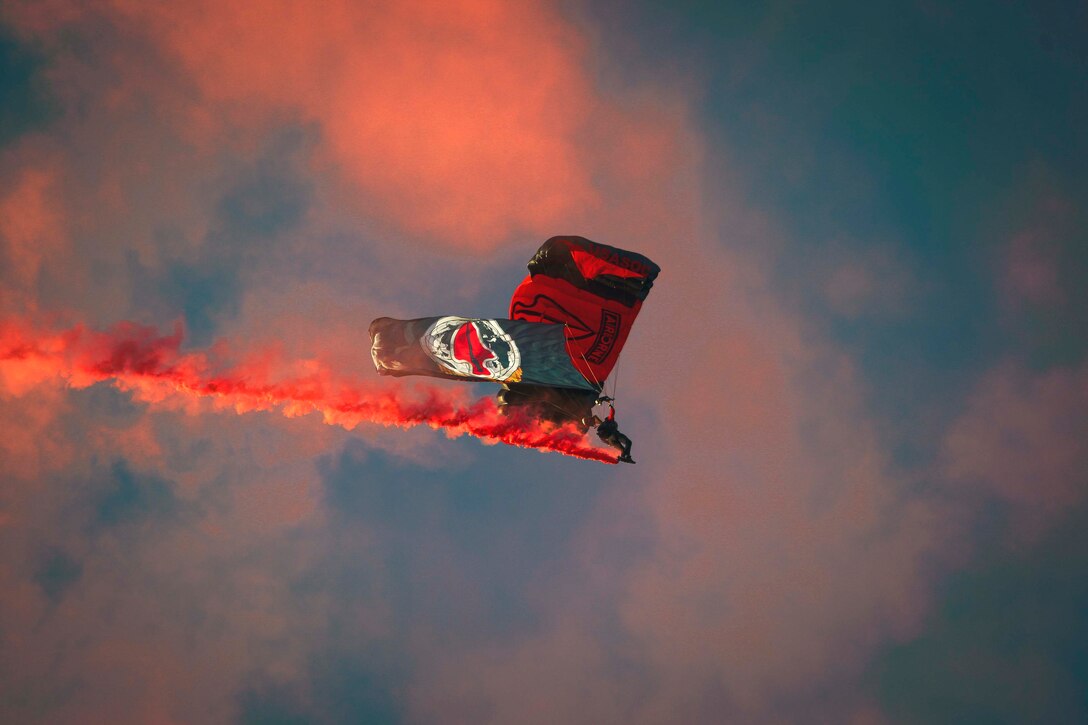 A soldier wearing a parachute descends in the sky with a flag as red smoke surrounds.