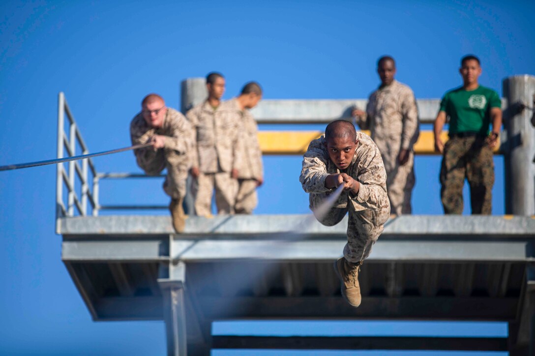 Two Marines Corps recruits slide along a rope suspended high above the ground as others watch and wait.