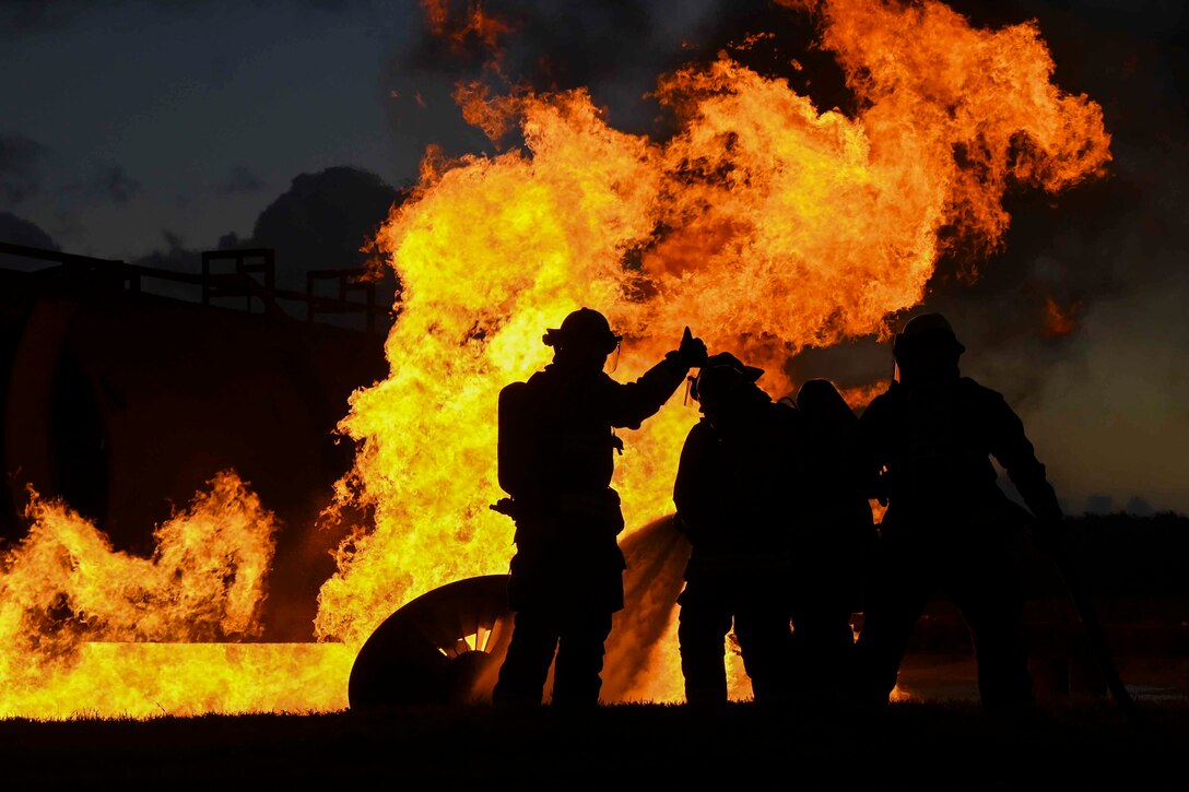 Airmen shown in silhouette stand in front of large flames.