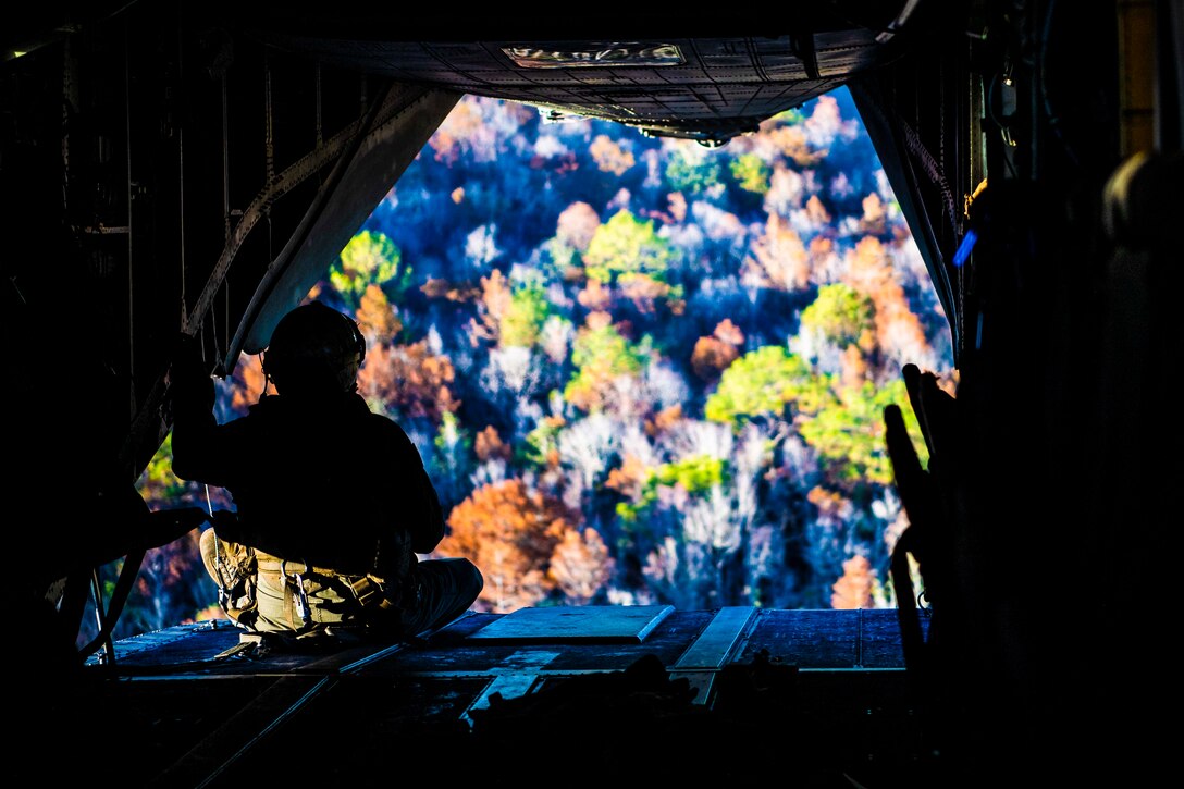 A Marine sits at the back of an aircraft looking at a tree line.