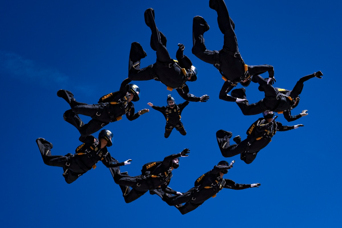 Army parachutists in black uniforms form a circle in a blue sky.