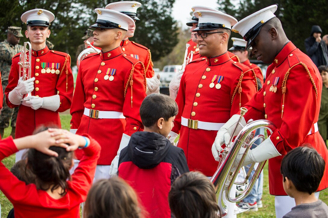 A group of Afghan children stand in front of a group of Marine Corps musicians in red uniforms.