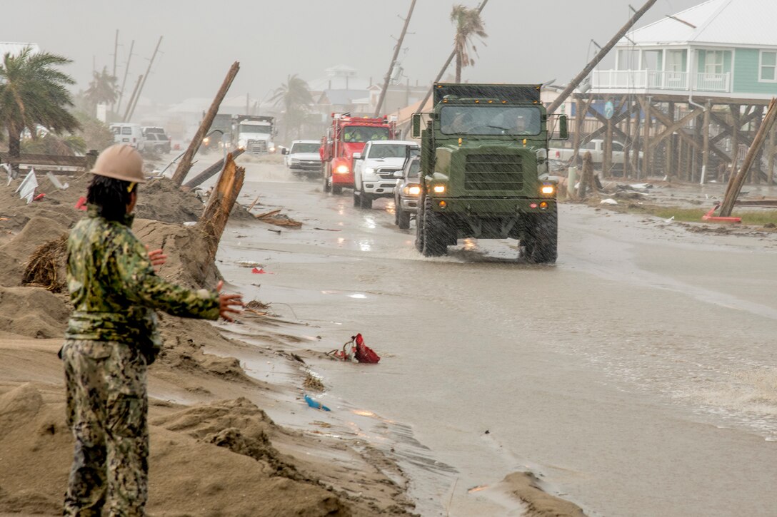 A sailor in a hard hat signals by the side of a road to a dump truck driving on it during a rainstorm.