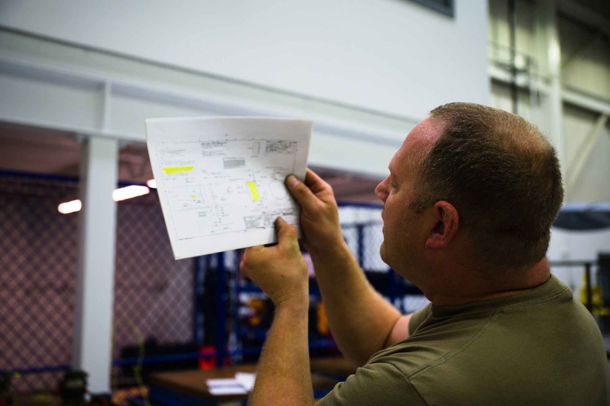 A man in a tan t-shirt holds up an engineering drawing of a bleed air shut-off "football" valve.
