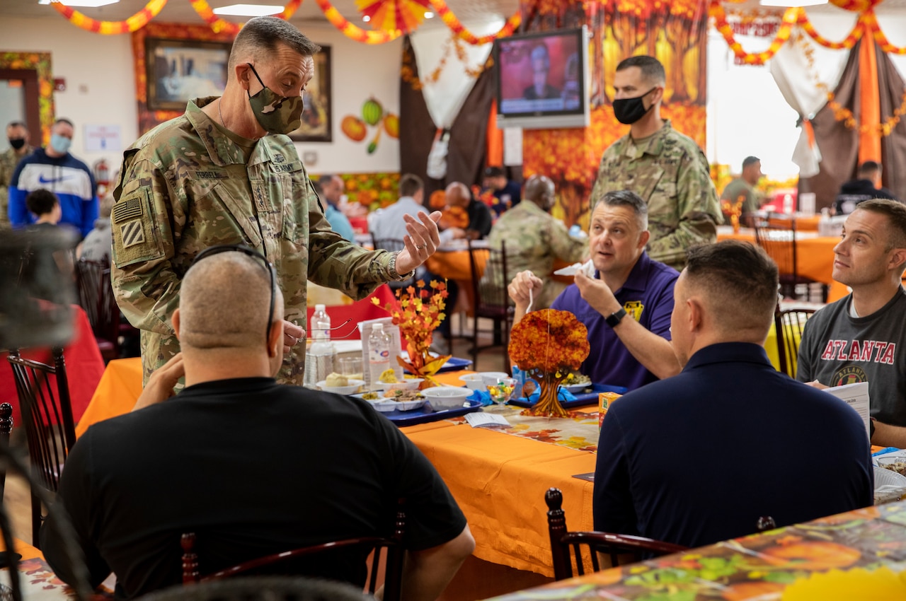 A soldier chats with other men seated at a table.