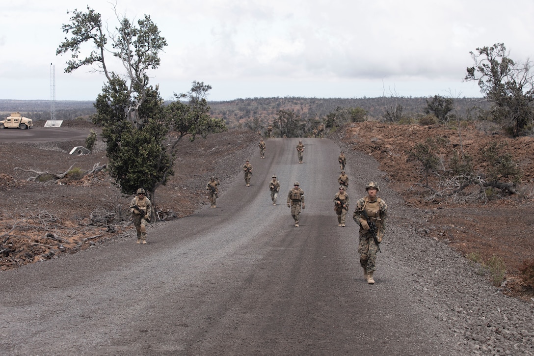 Marines carrying weapons walk on a dirt road.