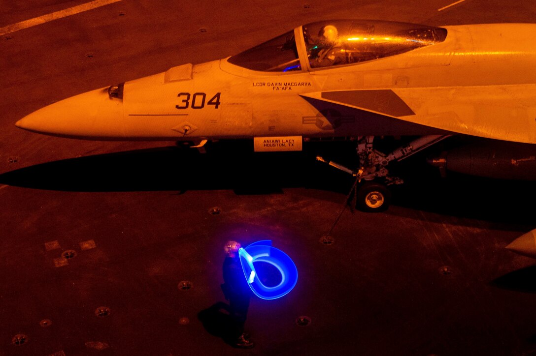 A sailor signals to an aircraft in the dark illuminated by a blue light.