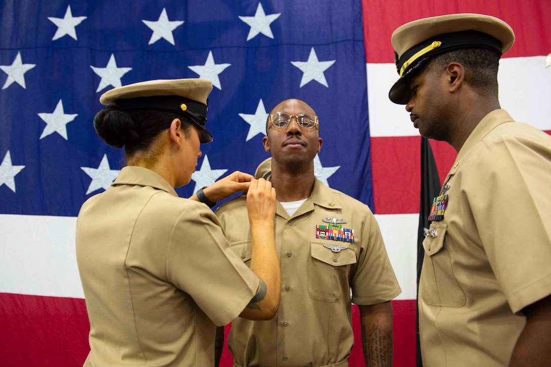 A sailor puts a pin on another sailor’s uniform as a different sailor stands beside them.