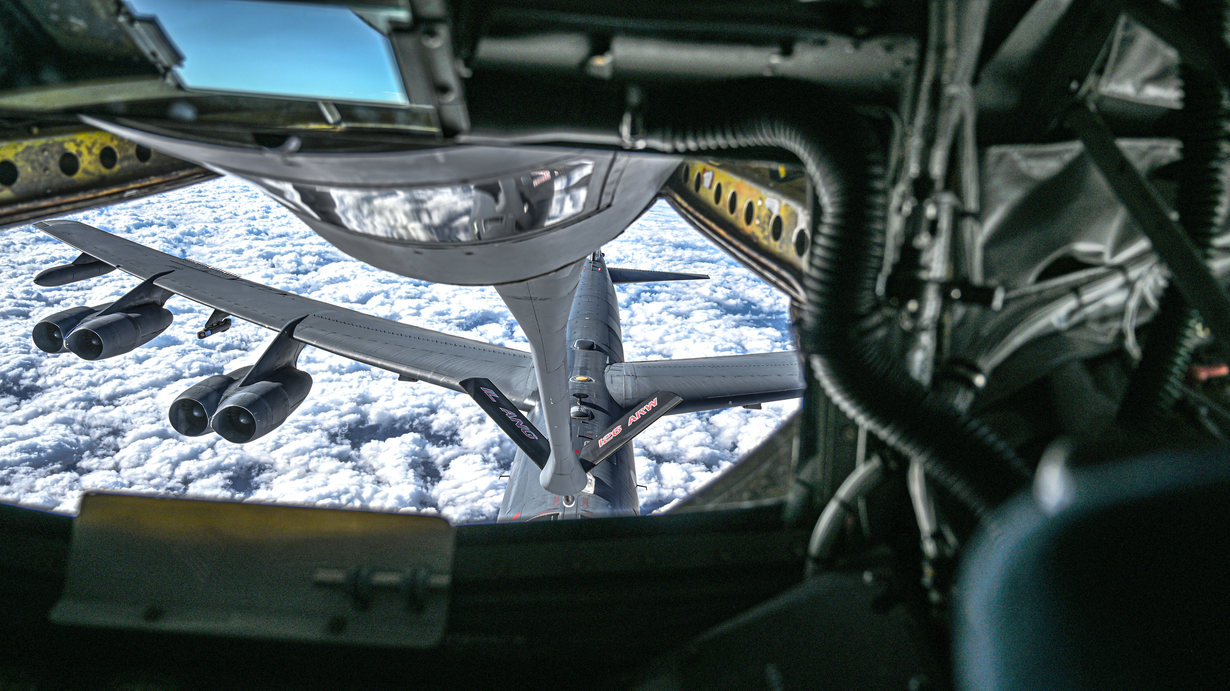 A wide angle look out the boom pod of a KC 135 on to a B-52 Stratofortress. On the right hand side of the window are wires and cables