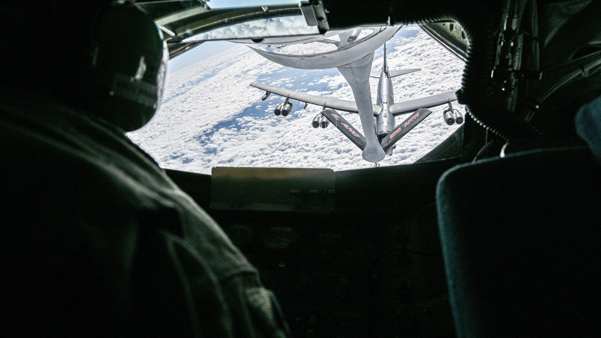 an airman looks out the back of a KC-135 looking at a B-52 bomber with the clouds below