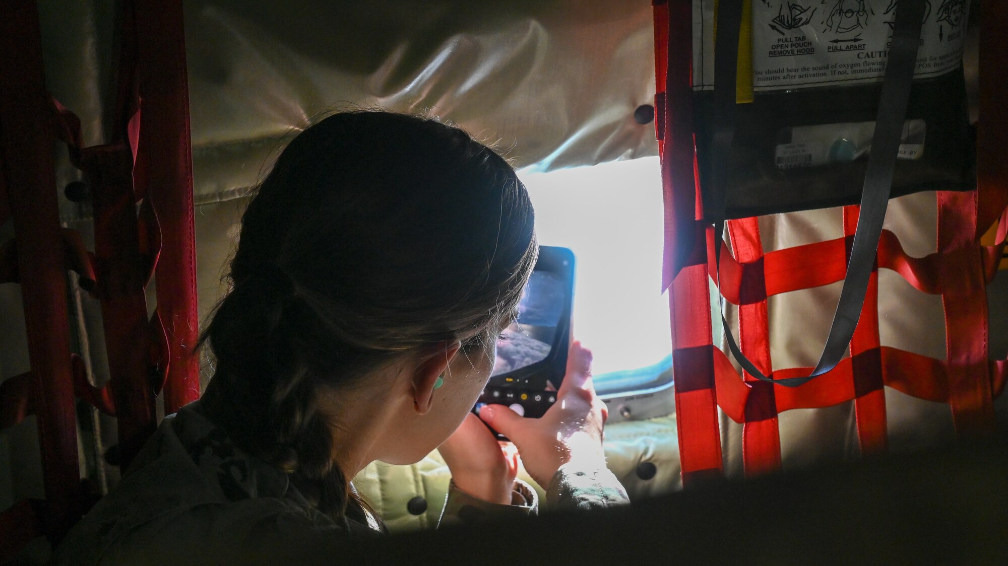 An airman looks out the leftside of a window of a KC-135 taking a photo with her phone.