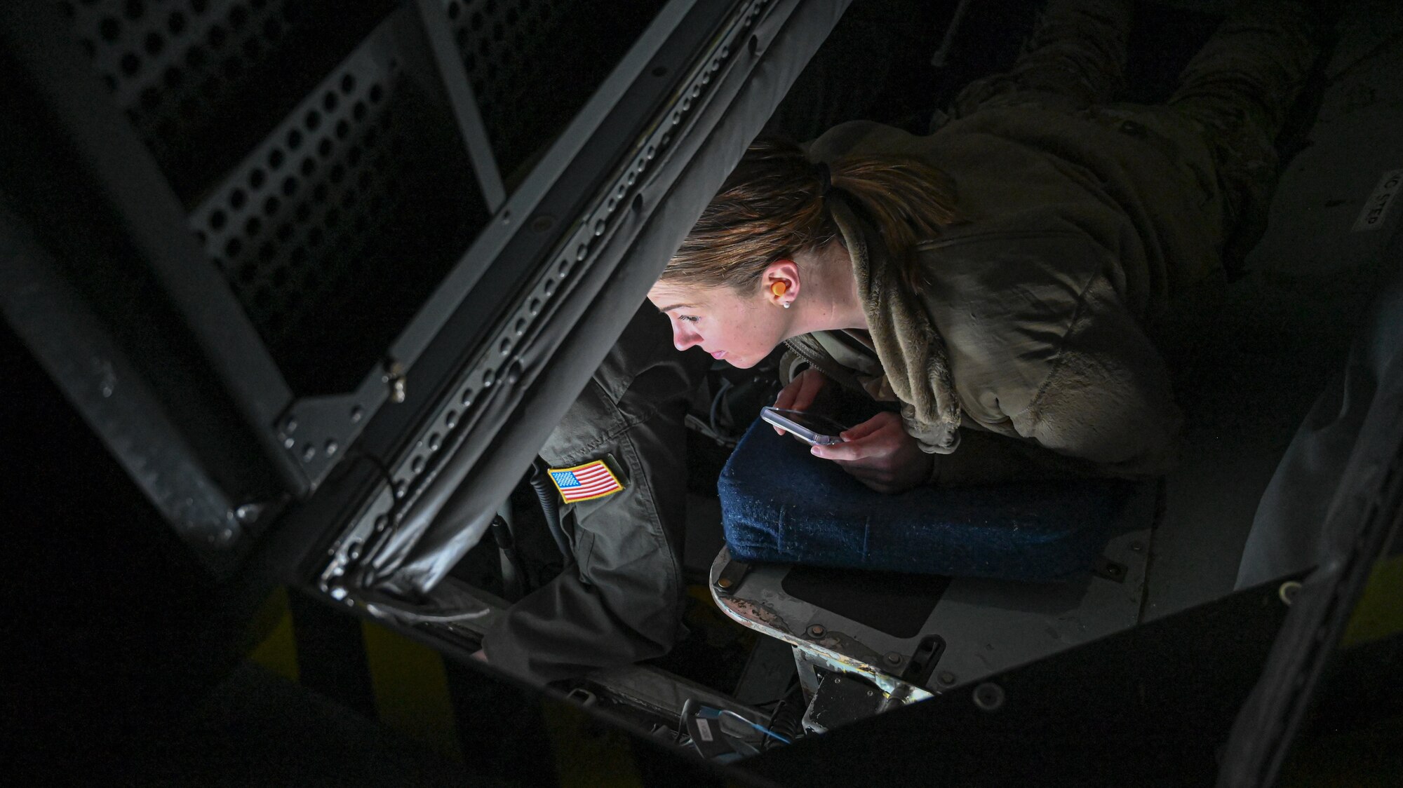 An airman laying down in a pod to the left of the boom operator looks through a window to see the plane being refueled