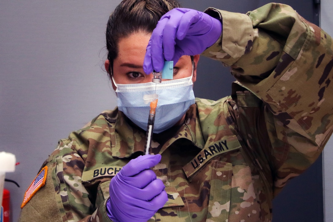 A soldier wearing a face mask looks at a syringe while inserting it into a vial.