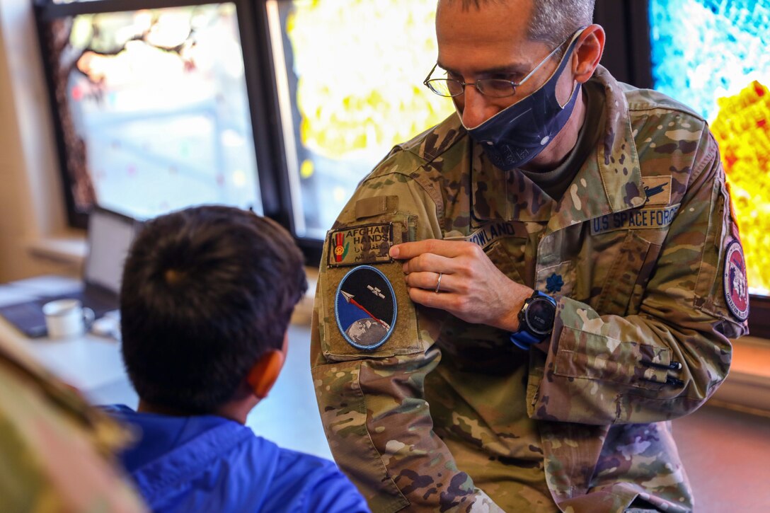 An Army soldier wearing a face mask points to a patch on his uniform sleeve while looking at a child.