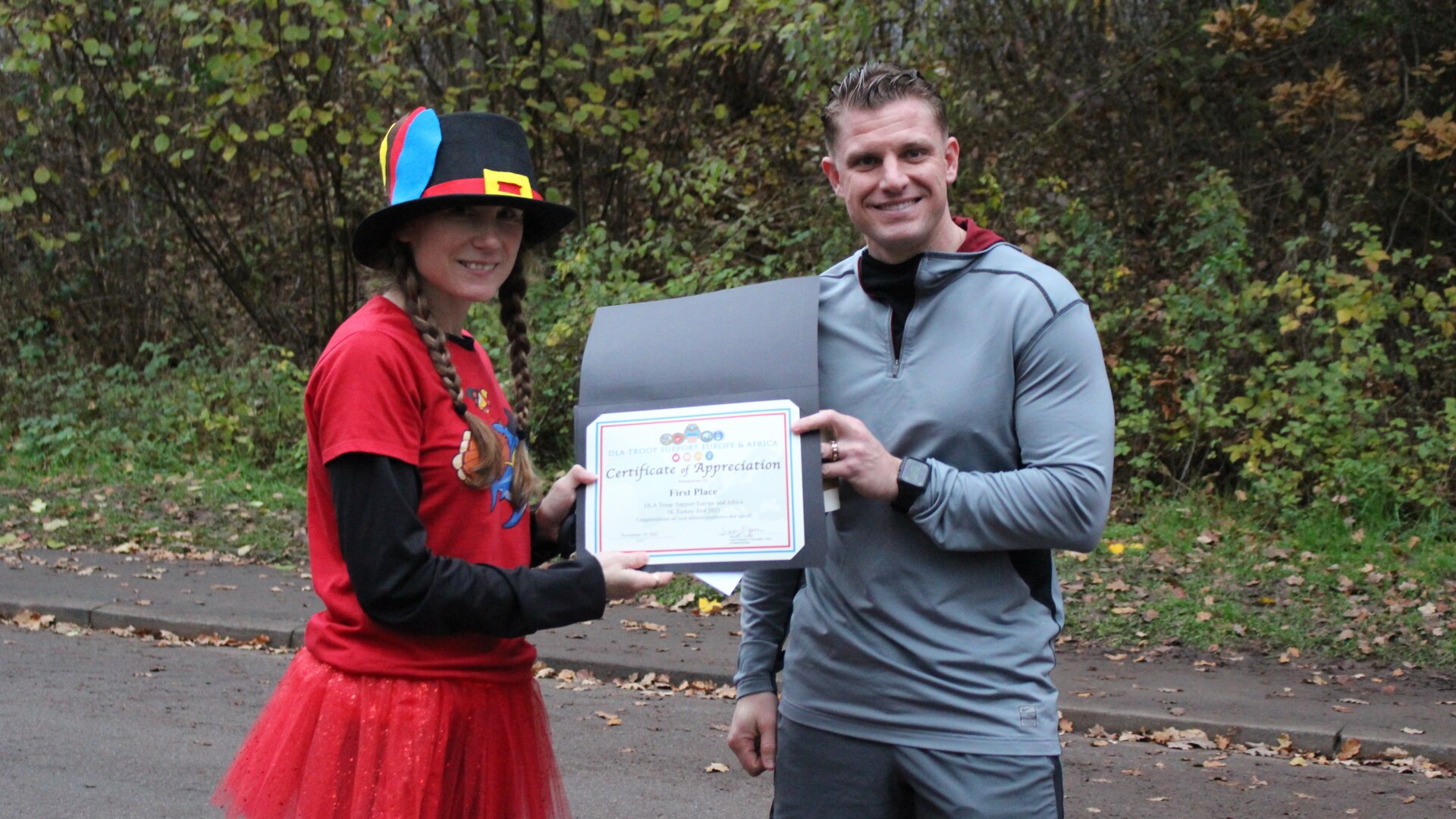 A woman in festive Thanksgiving run attire presents a certificate to a man in gray running gear.