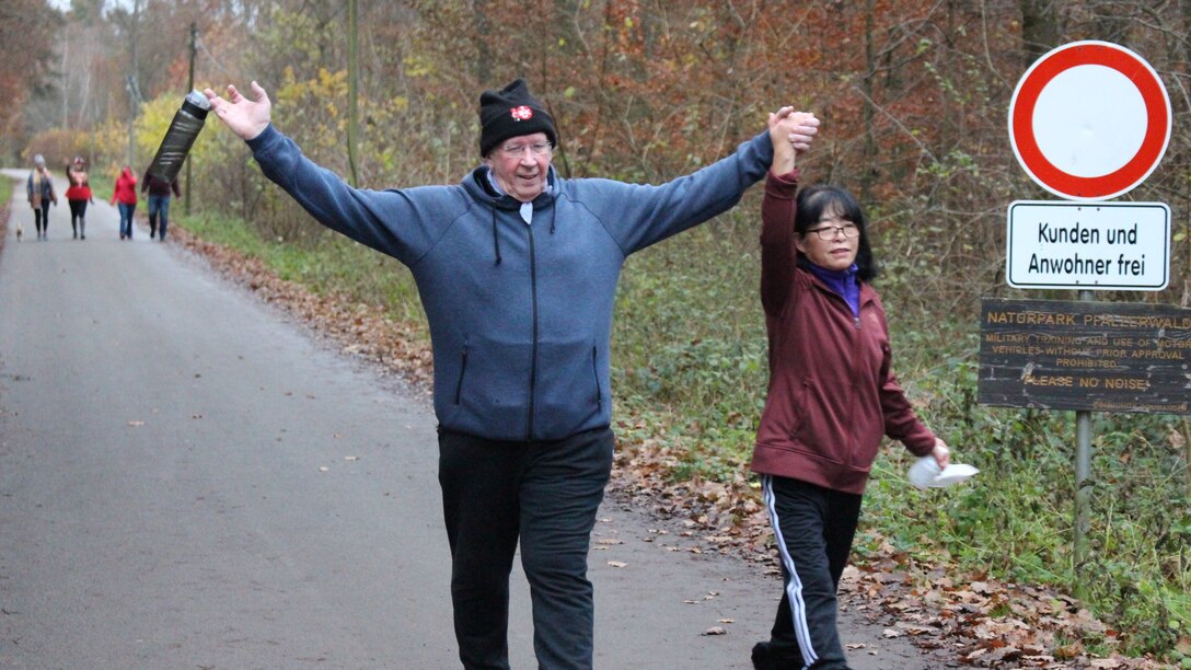A man raises his arms in celebration as he walks along holding a woman's hand on a wooded path.