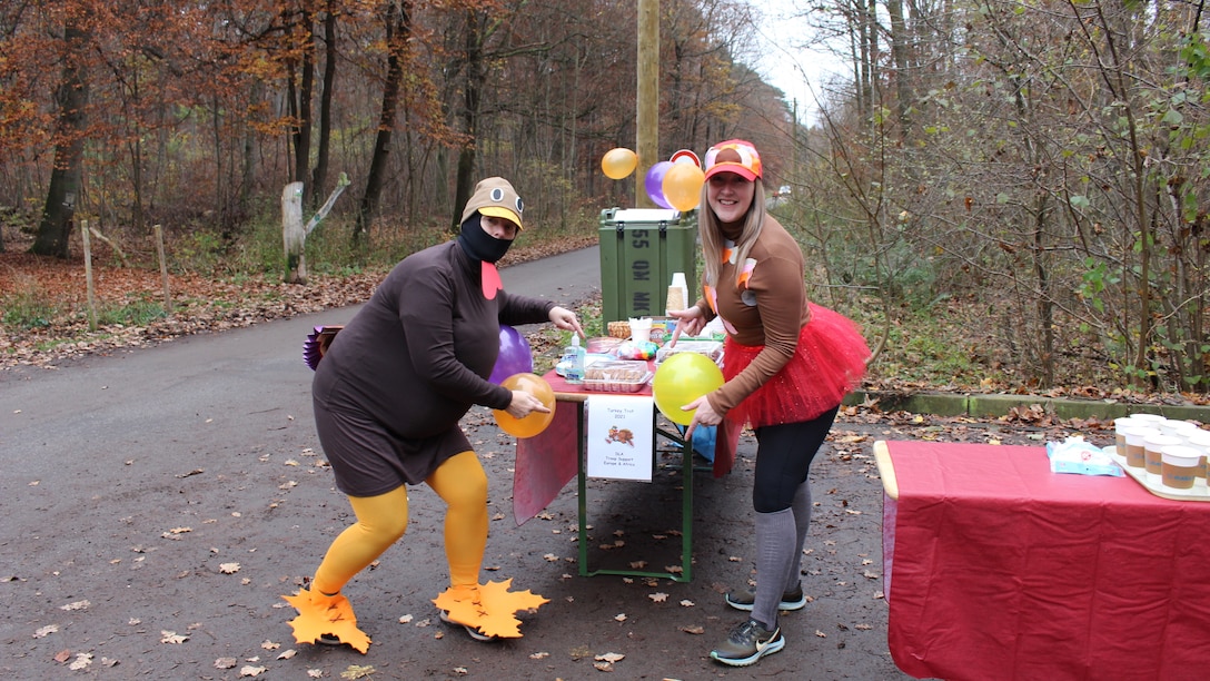 Two women in turkey-themed costumes stand on either side of a table with refreshments along a running path.