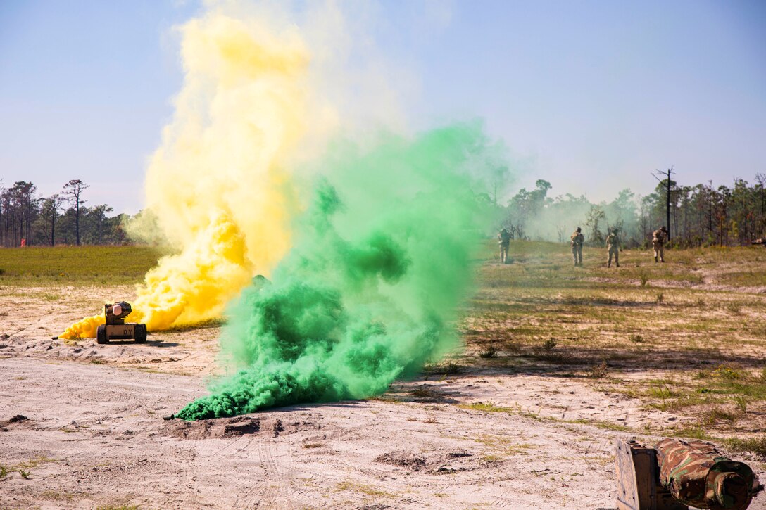 Marines train in a field while small vehicles drive around releasing green and yellow smoke.