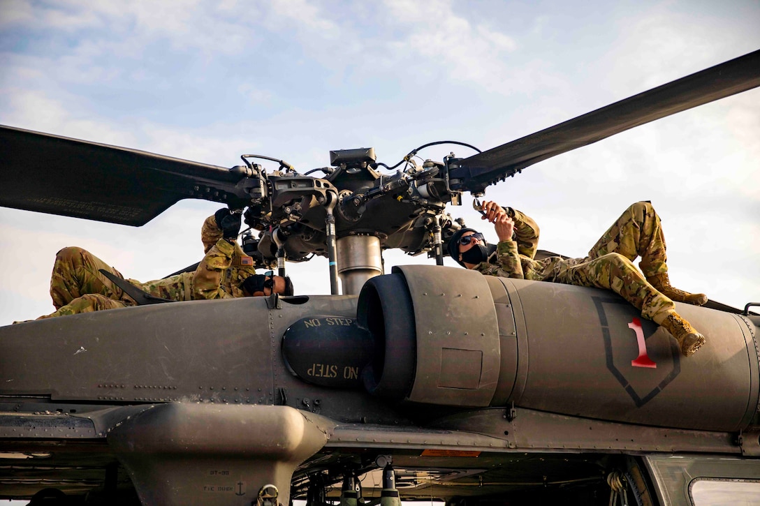 Two soldiers lie on top of a helicopter as they work on the blades.