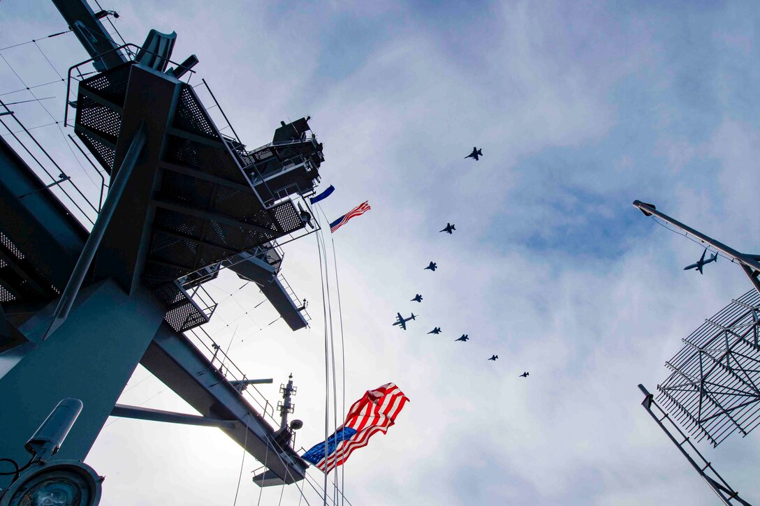 Nine aircraft fly in formation over a ship.