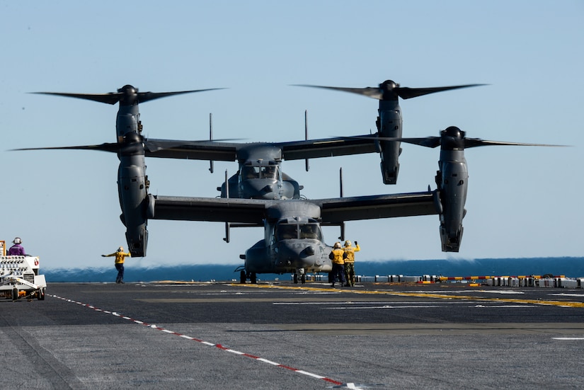 Two tilt-rotor aircraft hover over an aircraft carrier deck.