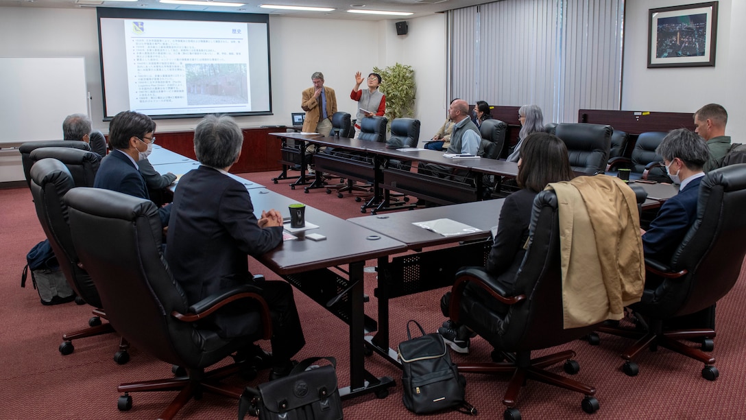 Air Force and Japanese officials sit at a large square table watch a slideshow in a conference room.
