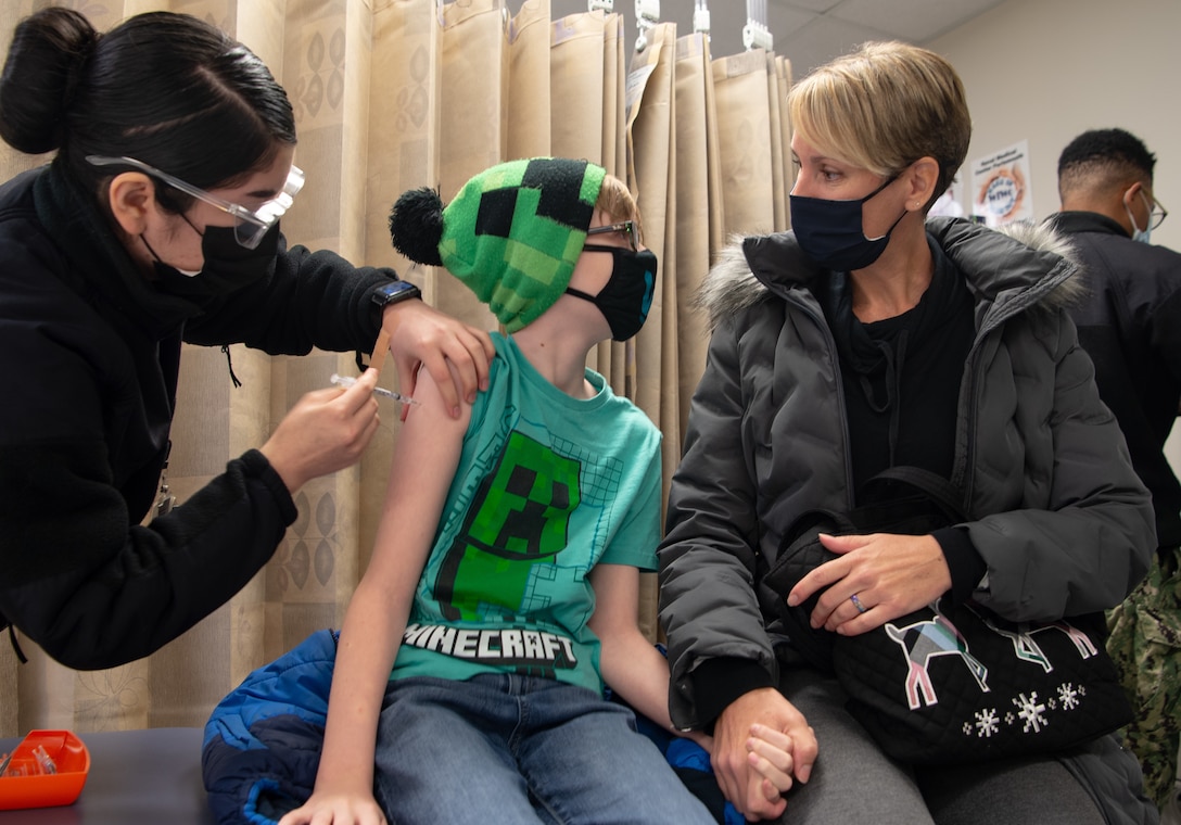 A child wearing a face mask holds hands and looks at a woman who is also wearing a face mask while a sailor administers a vaccine.